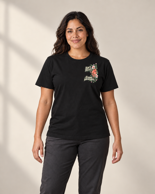 Woman with long dark wavy hair wearing the black Soft Heart Strong Boundaries T-shirt, floral heart graphic high on the wearer's left chest, front view.