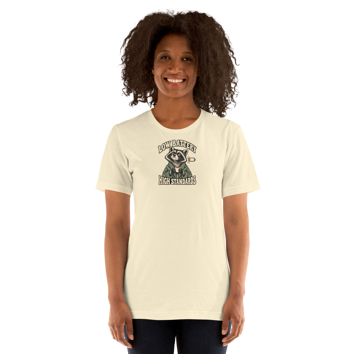 Woman modeling a natural-colored Low Battery High Standards t-shirt with a coffee-holding raccoon graphic on the front.